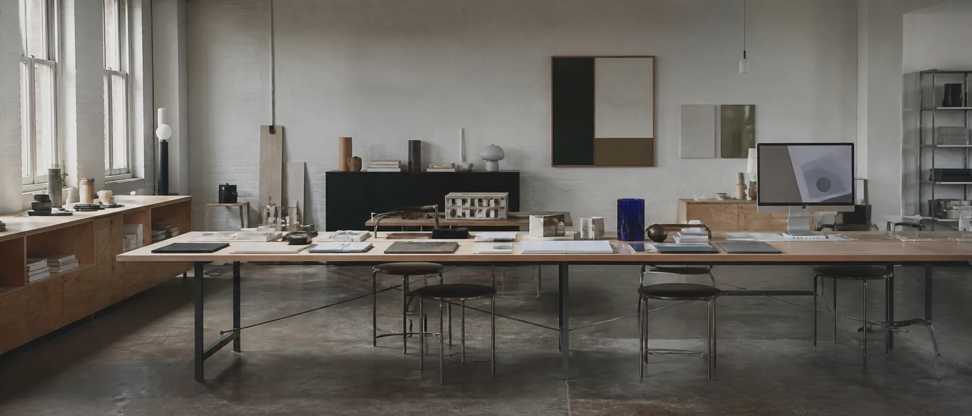 A long studio project table holds models, material samples and notebooks in soft daylight, with no people present.