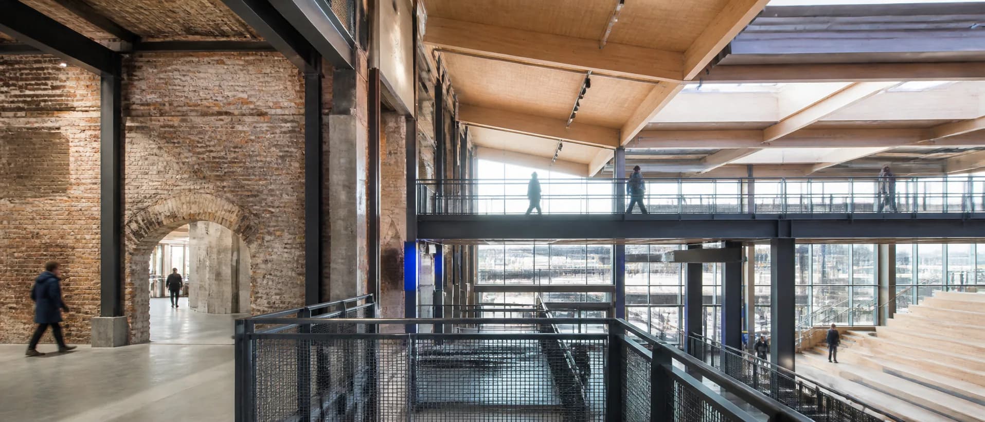 People move through a contemporary public interior where old brickwork meets new timber and steel structure.