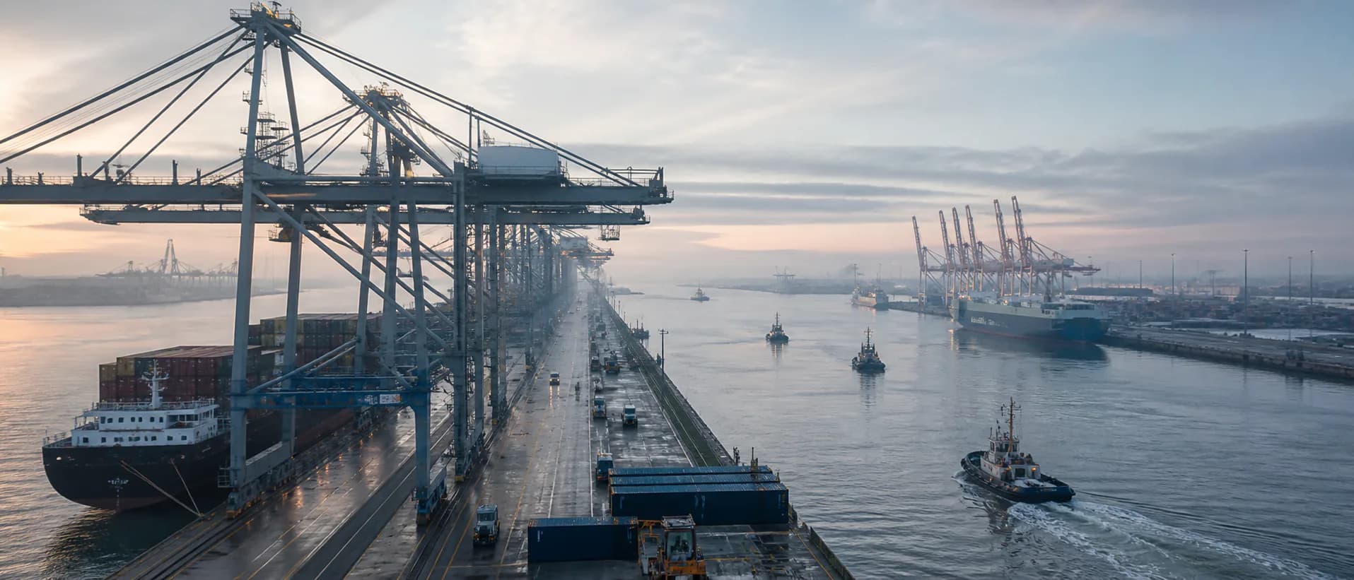 A wide harbour at dawn with quay cranes, service roads, vessels and tugboats moving through the channel.