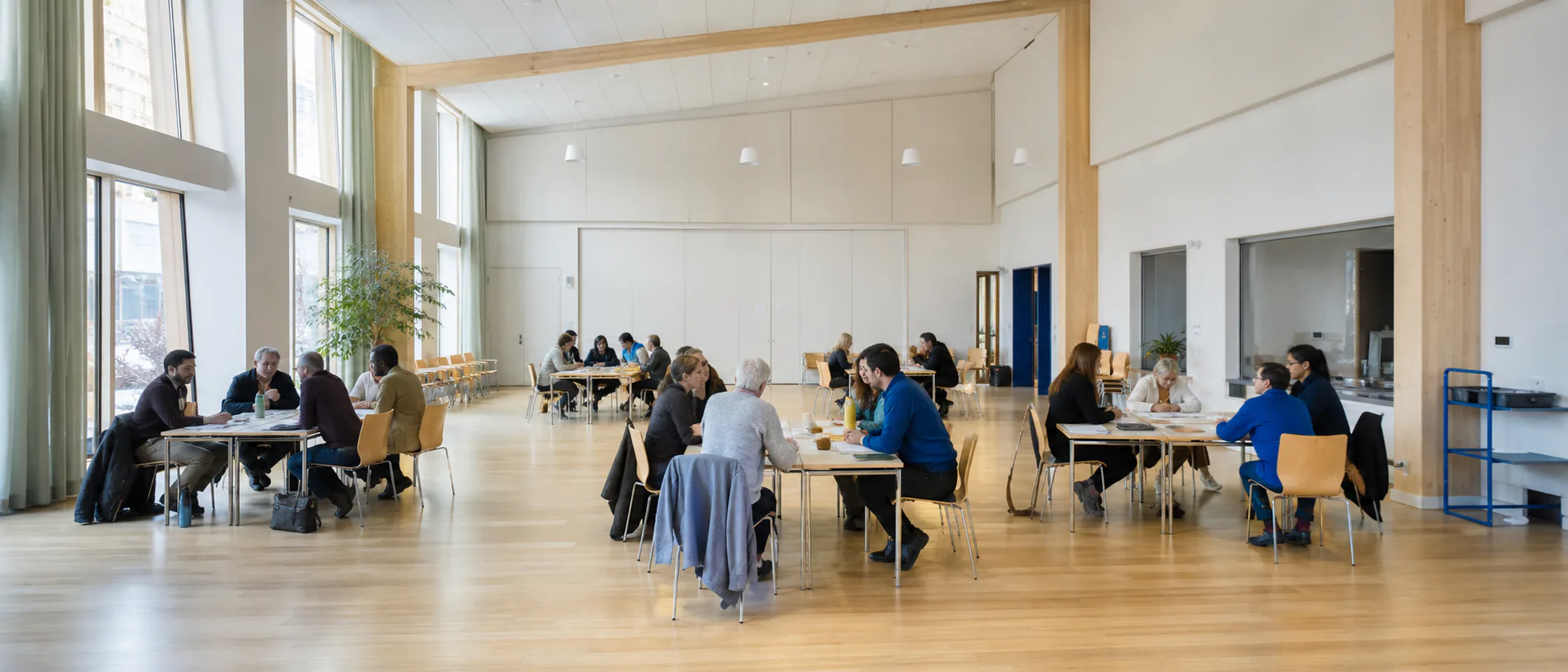 Adults work in small groups around tables in a bright, accessible community hall.